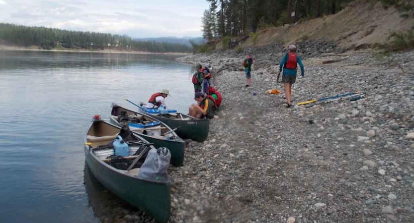 three canoes are beached on a shore of a calm lake. a few students carry gear away from the canoes. 